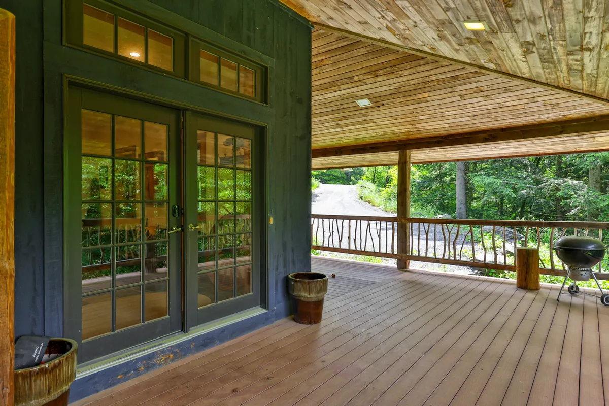 A cozy outdoor area featuring double glass doors leading from the home onto a porch with a wooden railing. A barbecue grill and comfortable seating are visible, with natural forest surroundings in the background. The scene is set for outdoor dining and social gatherings, with hints of sunset lighting.