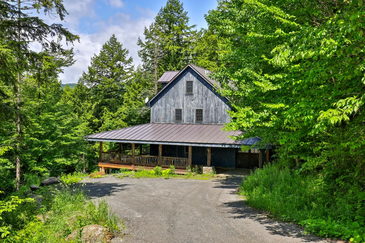 This image captures the rustic wooden house with its expansive porch wrapping around, set amidst lush greenery. The setting sun casts a warm glow, highlighting spaces for relaxing, storytelling by a fire pit, and direct access to nature trails just steps away.
