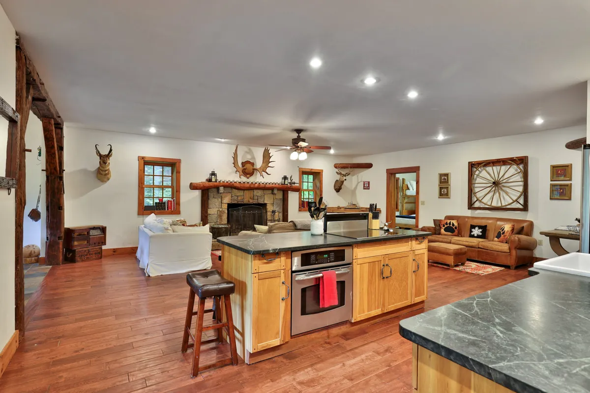 A spacious open-concept area that seamlessly blends the kitchen, a cozy seating space with plush leather sofas, and a prominent stone fireplace. The kitchen features a central island and high-end appliances, while the living space is arranged for group relaxation and warmth by the fire, all unified by rustic decor and inviting lighting.