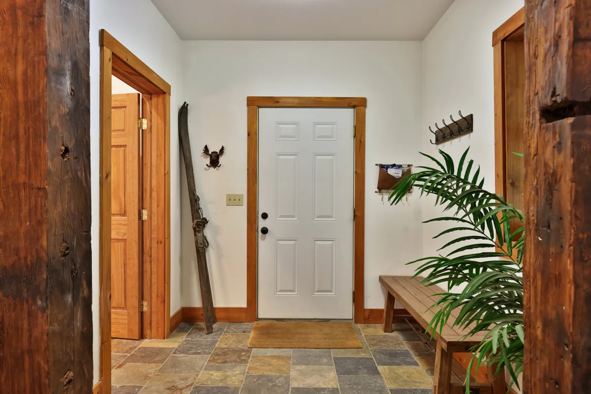 A view of the lodge's welcoming front entryway featuring a sturdy wooden front door, a cozy wooden bench, decorative wall art, a potted plant, and a rustic stone floor. The space is designed to greet guests warmly as they arrive.