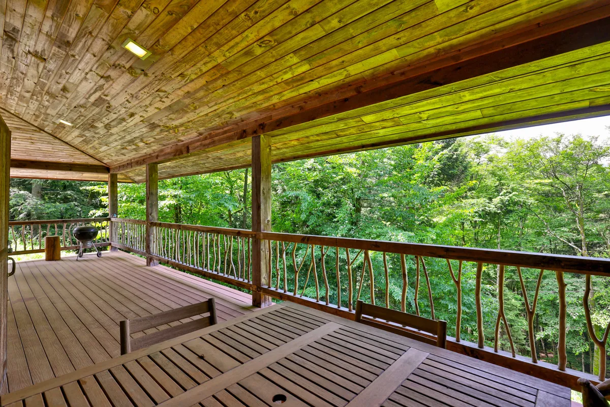 A wooden deck surrounded by trees, featuring an outdoor dining table and a grill. The inviting setting is perfect for enjoying meals outside, relaxing in nature, or hosting gatherings, with forest views in the background.