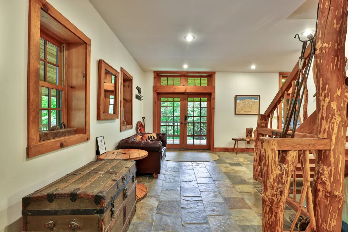 A sun-filled communal living space with a leather couch, vintage trunk, wooden staircase, decorative frames, and glass doors leading toward the outdoors. The room exudes comfort and encourages gathering and relaxation.