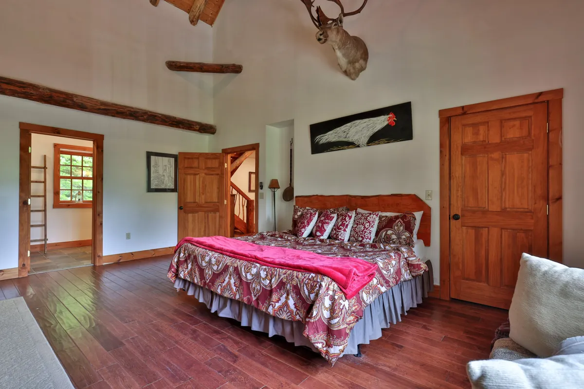 A warm and inviting bedroom with a large bed adorned in ornate bedding, a mounted deer head above the bed, wooden doors, and artwork on the wall. The atmosphere is cozy and classic, capturing the essence of Vermont style and comfort.