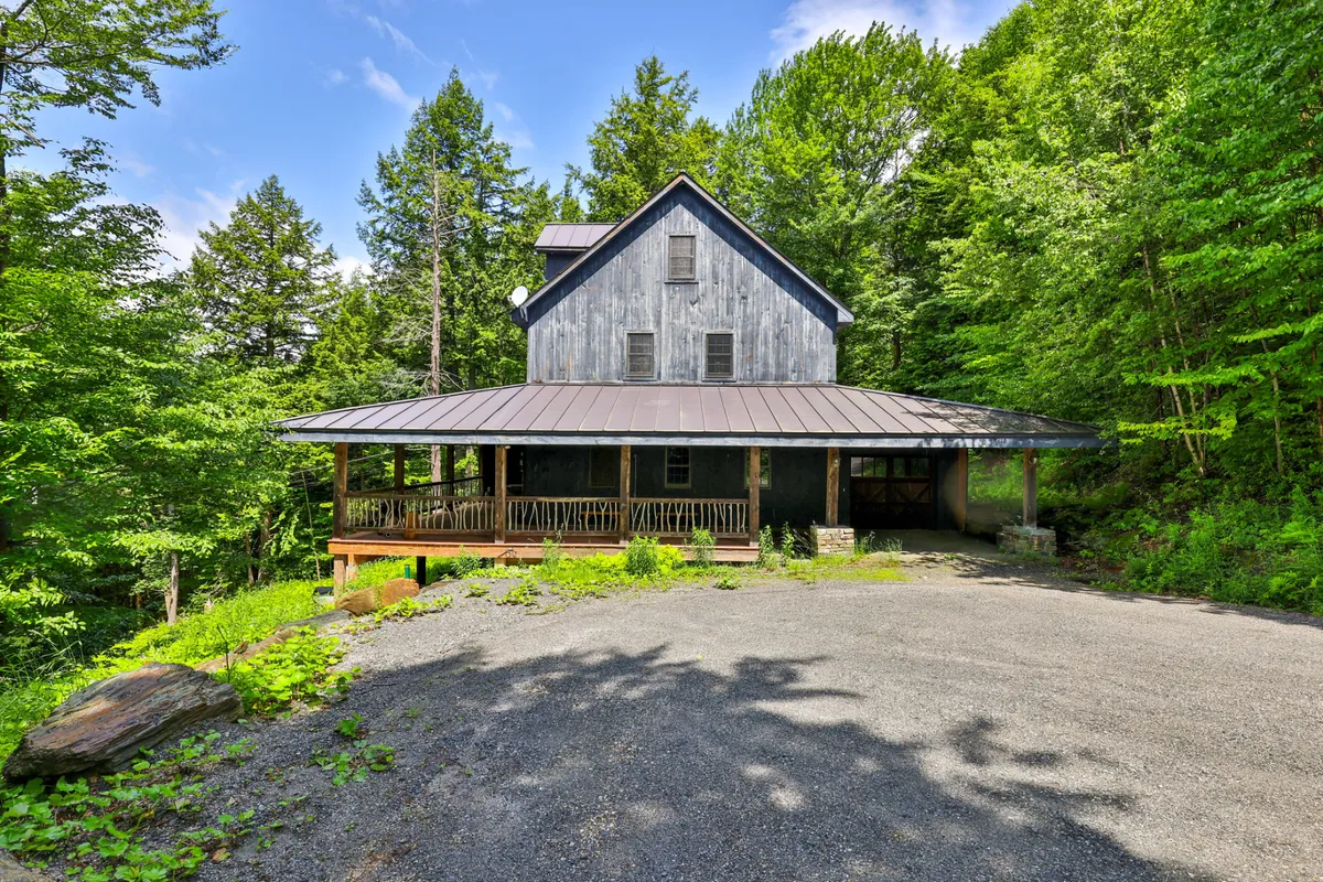 This image shows a broad wooden porch with a sturdy metal roof, surrounded by lush Vermont greenery. The porch is furnished for comfort, inviting relaxation while offering unobstructed panoramic views of the mountains. Early morning mist can be seen enveloping the landscape, creating a peaceful and immersive outdoor experience.