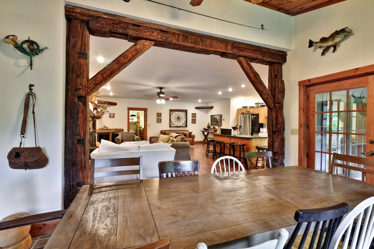A spacious interior view featuring a large wooden dining table with ample seating, exposed beams, and an adjacent comfortable seating area. The kitchen is partially visible, emphasizing opportunities for group cooking and shared meals.
